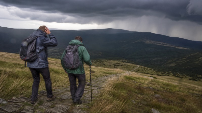 Tmavá bouřková oblaka nad skalnatým horským hřebenem, po pěšině pod hřebenem sestupuje turista v pláštěnce