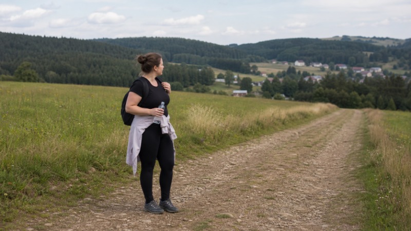 Turista s malým batohem stojí na lesní pěšině u stromu s turistickou značkou a dívá se do mapy