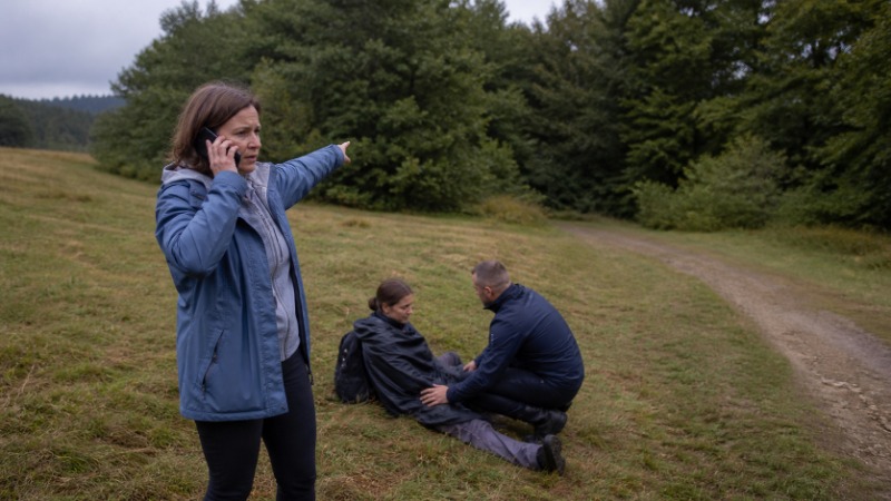 Turista v bundě stojí v mlze na kamenitém hřebeni u rozcestníku a drží mobilní telefon, vedle něj leží odložený batoh